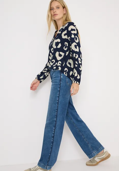Frau mit gemustertem Pullover, weiter Jeans und Sneakers geht vor weißem Hintergrund.